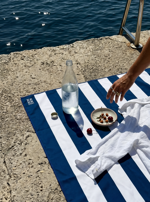 Amalfi lightweight microfibre travel towel with blue and white stripes by the water as a woman reaches for a bowl of cherries