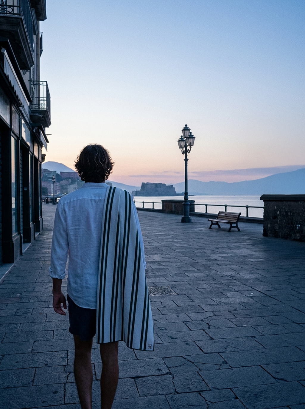 Man carrying the Positano compact microfibre travel towel with white and dark green stripes over his shoulder by the waterfront at blue hour