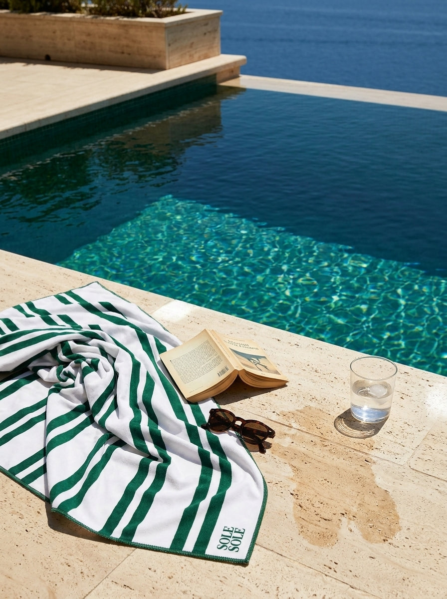 Positano quick-dry microfibre travel towel with white and dark green stripes beside a pool with a book, sunglasses, and a glass of water