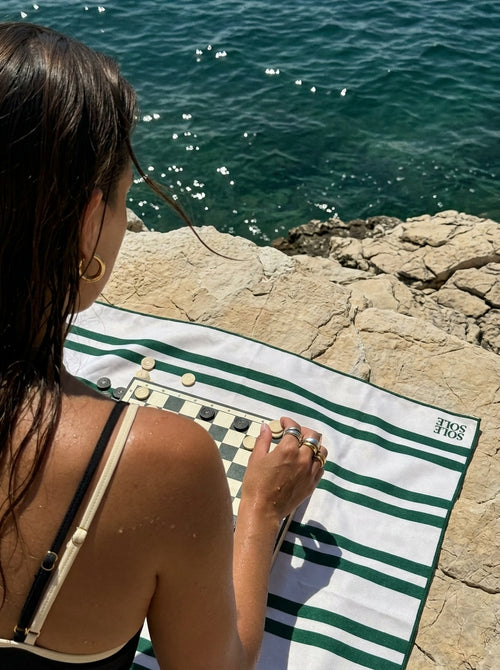 Woman playing a board game on the Positano lightweight microfibre travel towel with white and dark green stripes beside a coastal cliff